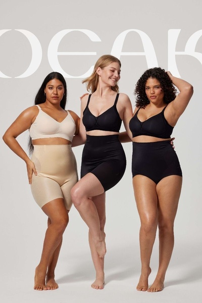 Three models of varying body types pose together in neutral and black shapewear sets against a minimal, soft-toned background.